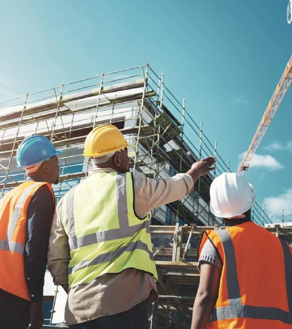 Three site workers looking up at a building under construction, with scaffolding and a tower crane behind it.